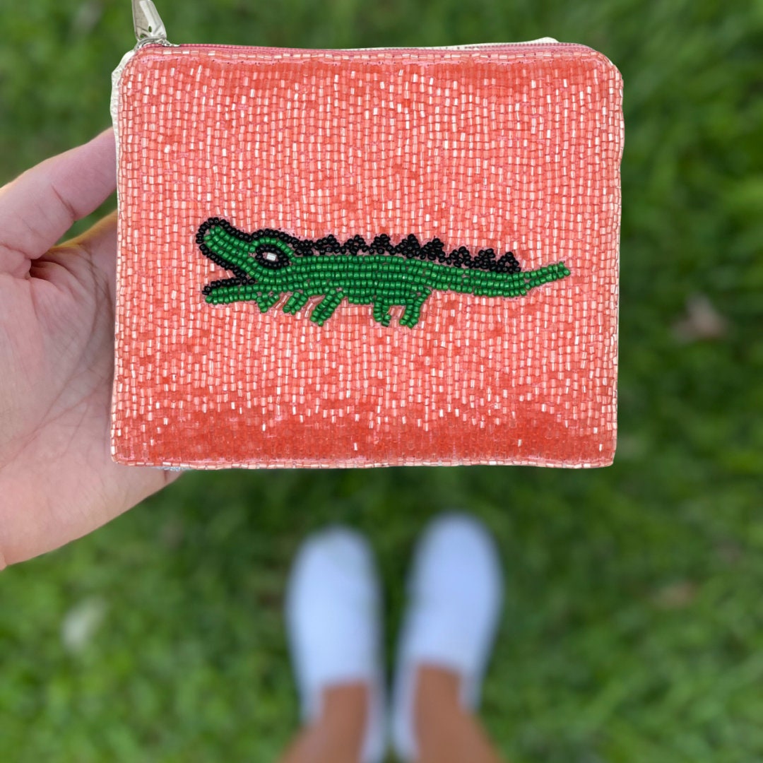 Hand holding a small pink pouch with a green alligator design against a grass background