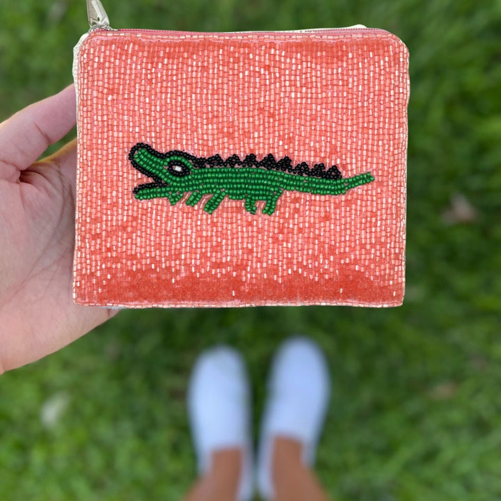 Hand holding a small pink pouch with a green alligator design against a grass background